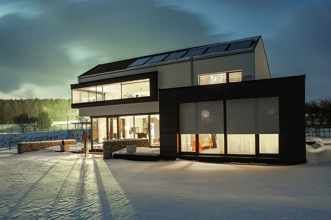 Außenansicht eines modernen 2-geschossigen Wohnhauses im verschneiten Garten, mit hell erleuchteten Fenstern und halb geschlossenen Rollladen