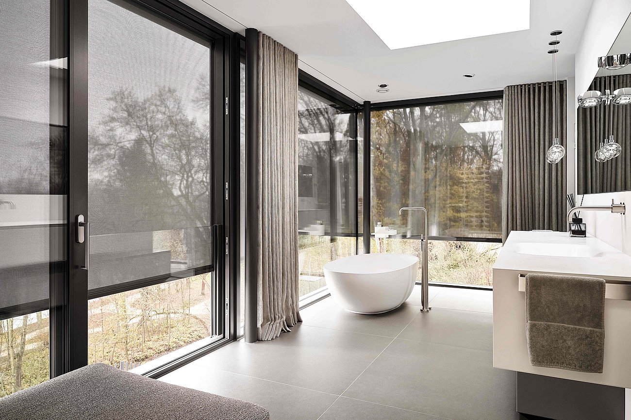 Innenaufnahme eines modern eingerichteten Badezimmers mit freistehender Badewanne, verglaster Zimmerecke und halb geschlossenen Textilscreens
